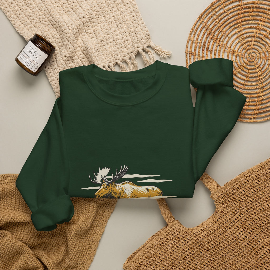 Forest Green North Woods Moose Crewneck laid flat next to a woven basket and knit blanket