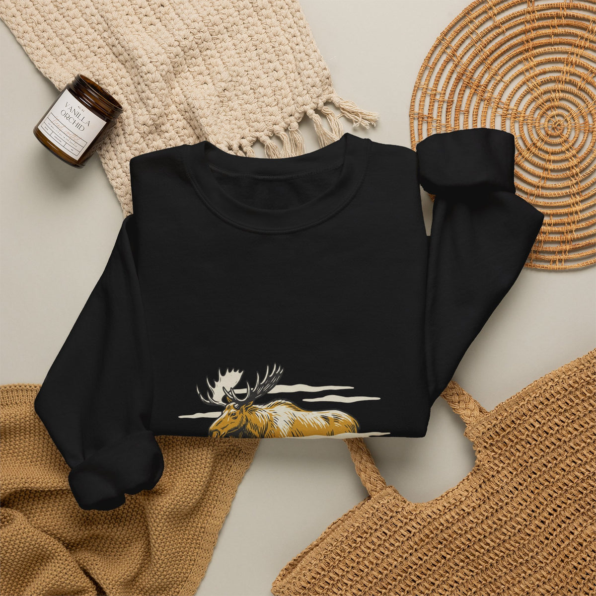 Black North Woods Moose Crewneck laid flat next to a woven basket and knit blanket